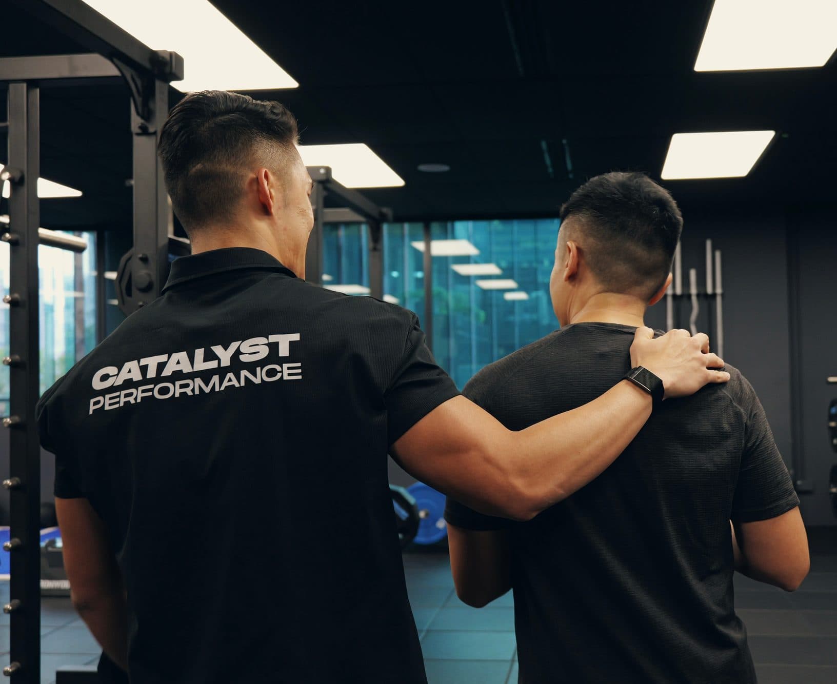 Catalyst Performance coach in a branded polo with an arm around a male member at the rack, Singapore CBD personal training studio