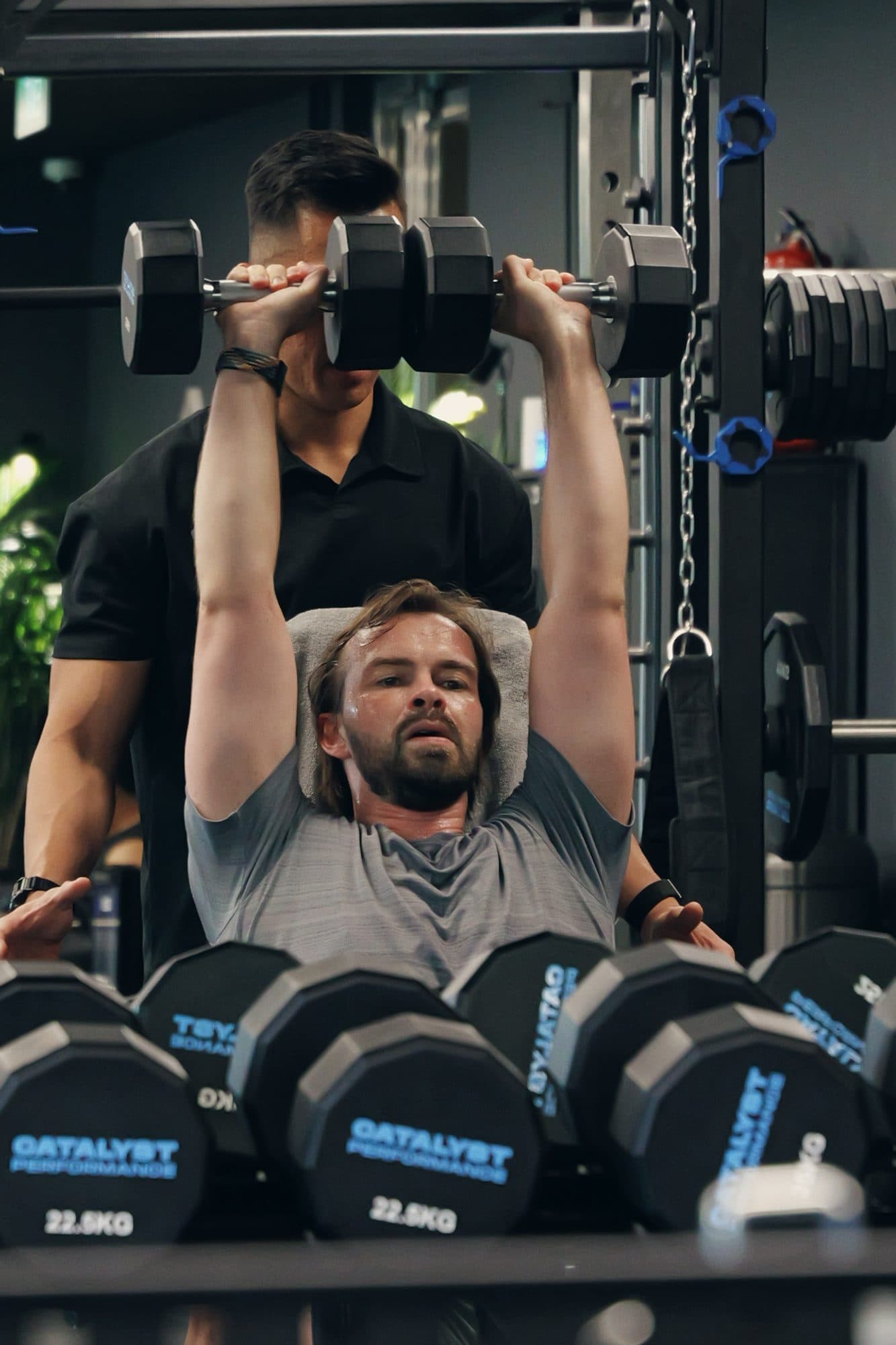 Catalyst Performance member pressing dumbbells with a coach spotting, Catalyst-branded plates in frame, Singapore CBD strength coaching