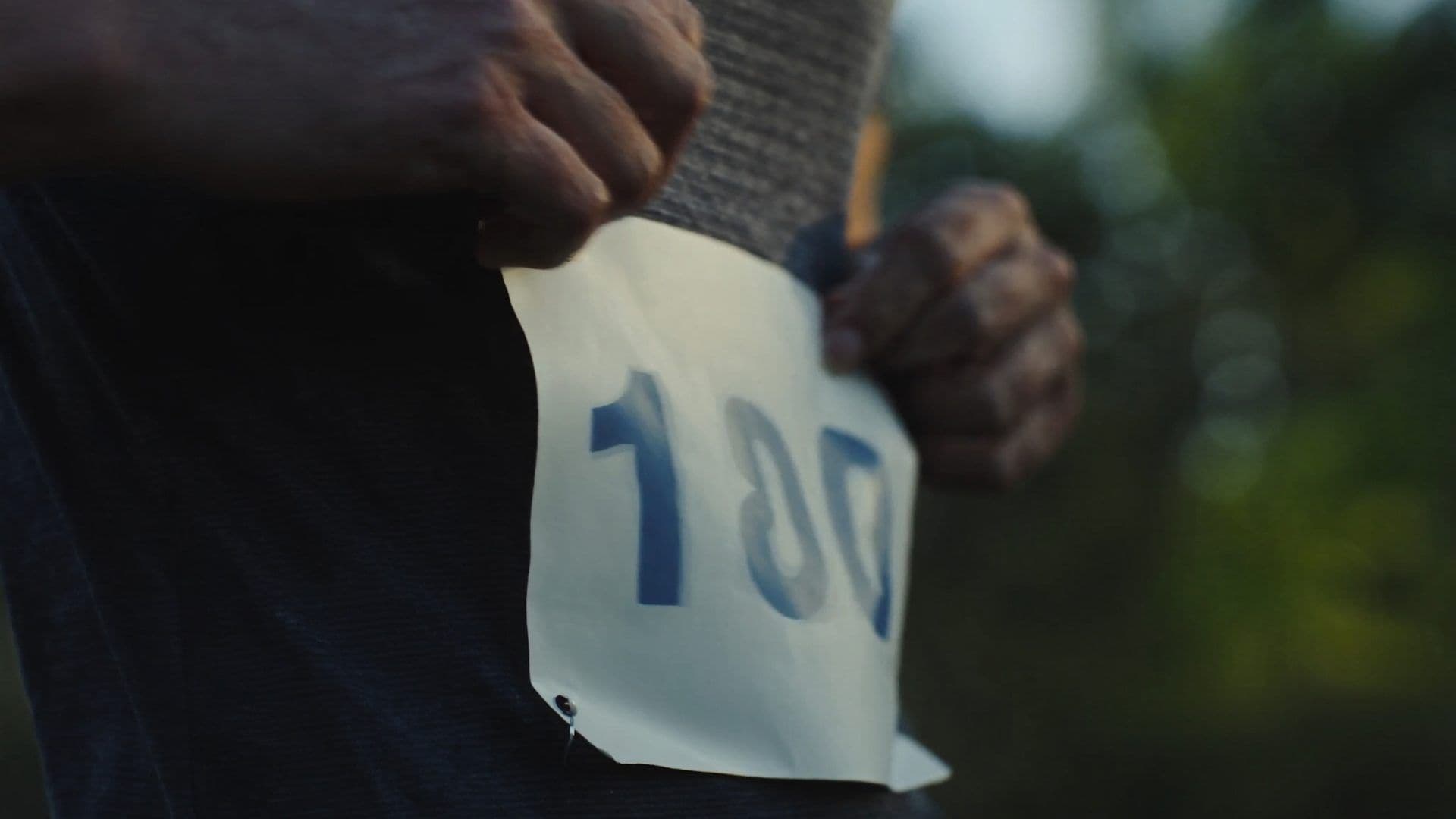 A runner's hand fastening a race bib at the start line, charcoal and steel-blue editorial palette, close-up tropical morning light