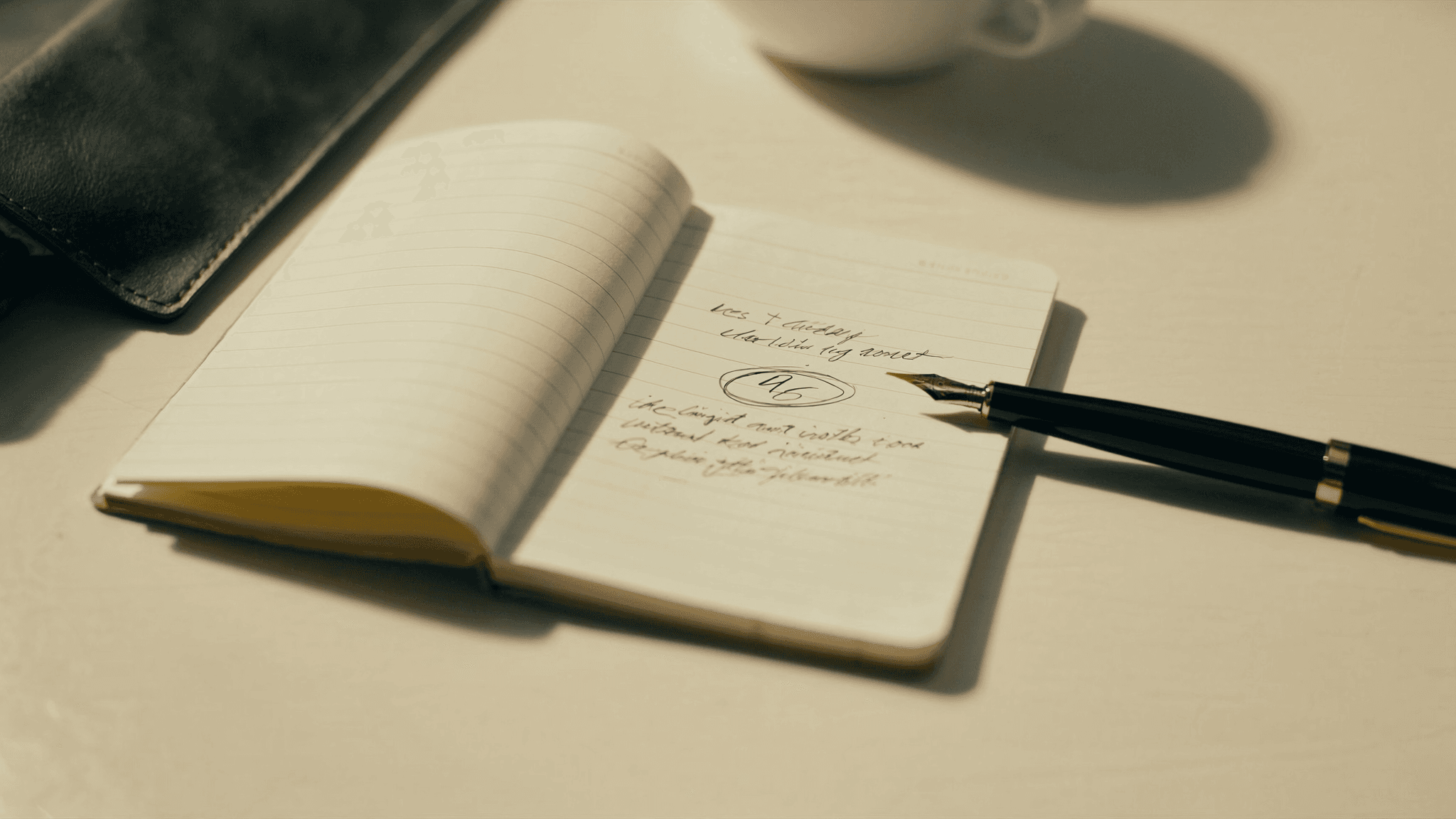 A small ruled notebook open on a warm cream wooden desk, a single fountain-pen-written number circled in black ink on the right-hand page, with a polished black-and-brass fountain pen resting across the lower right corner; a charcoal leather portfolio and a small white ceramic coffee cup in soft focus background.