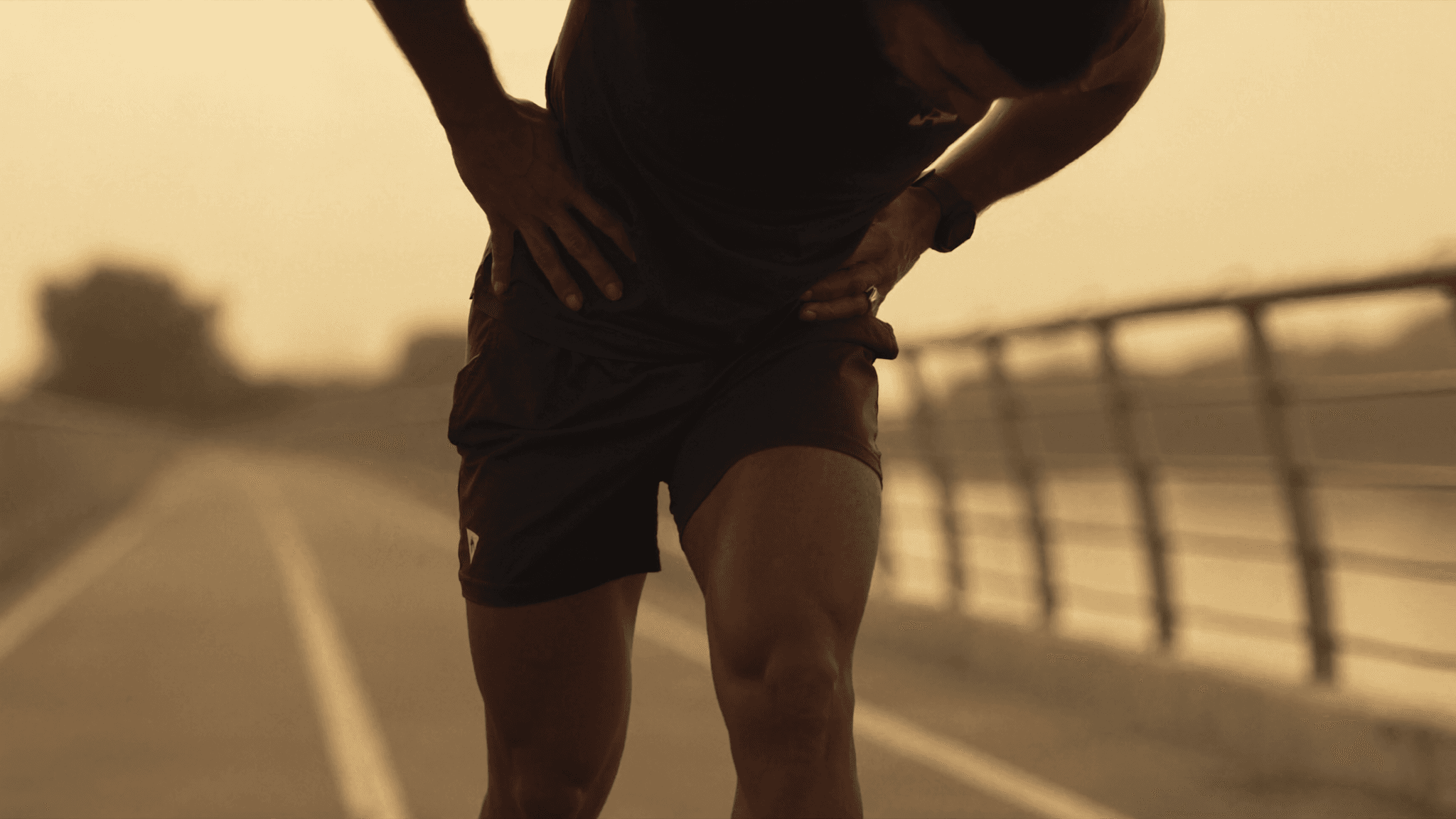 A runner pausing mid-stride on an East Coast Park style waterfront path, hands on hips, looking down at the right knee with mild discomfort. Side angle, golden hour Singapore light, no facial features visible, athletic clothing in neutral colours.