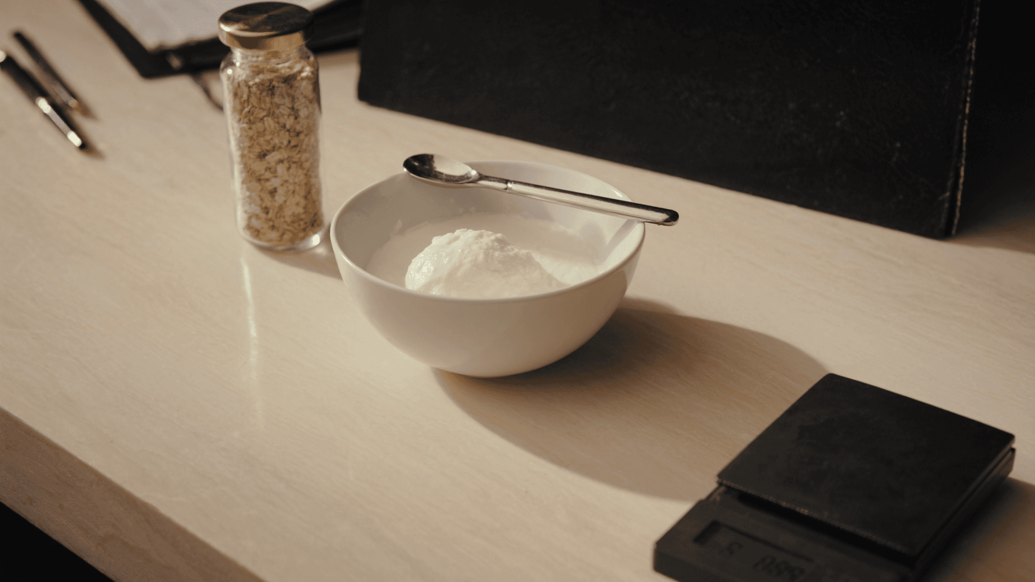 A quiet morning kitchen counter on a warm cream surface: a clean white ceramic bowl with portioned Greek yoghurt and a matte stainless steel spoon, a glass jar of rolled oats with a brass lid, a precision kitchen scale in matte charcoal partially in soft focus, and a black leather notebook in deeper soft-focus background.