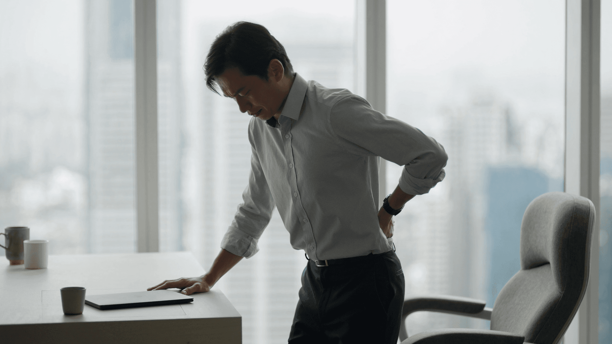 A late-30s Asian male in a smart-casual white button-down shirt stands beside a clean wooden desk in a minimalist Singapore CBD office. Side angle, one hand pressed firmly to his lower back, head slightly bowed forward in evident discomfort. A closed laptop and small ceramic cup sit on the desk; an ergonomic chair is pushed back. A floor-to-ceiling window behind him reveals the Singapore CBD skyline in soft daylight.