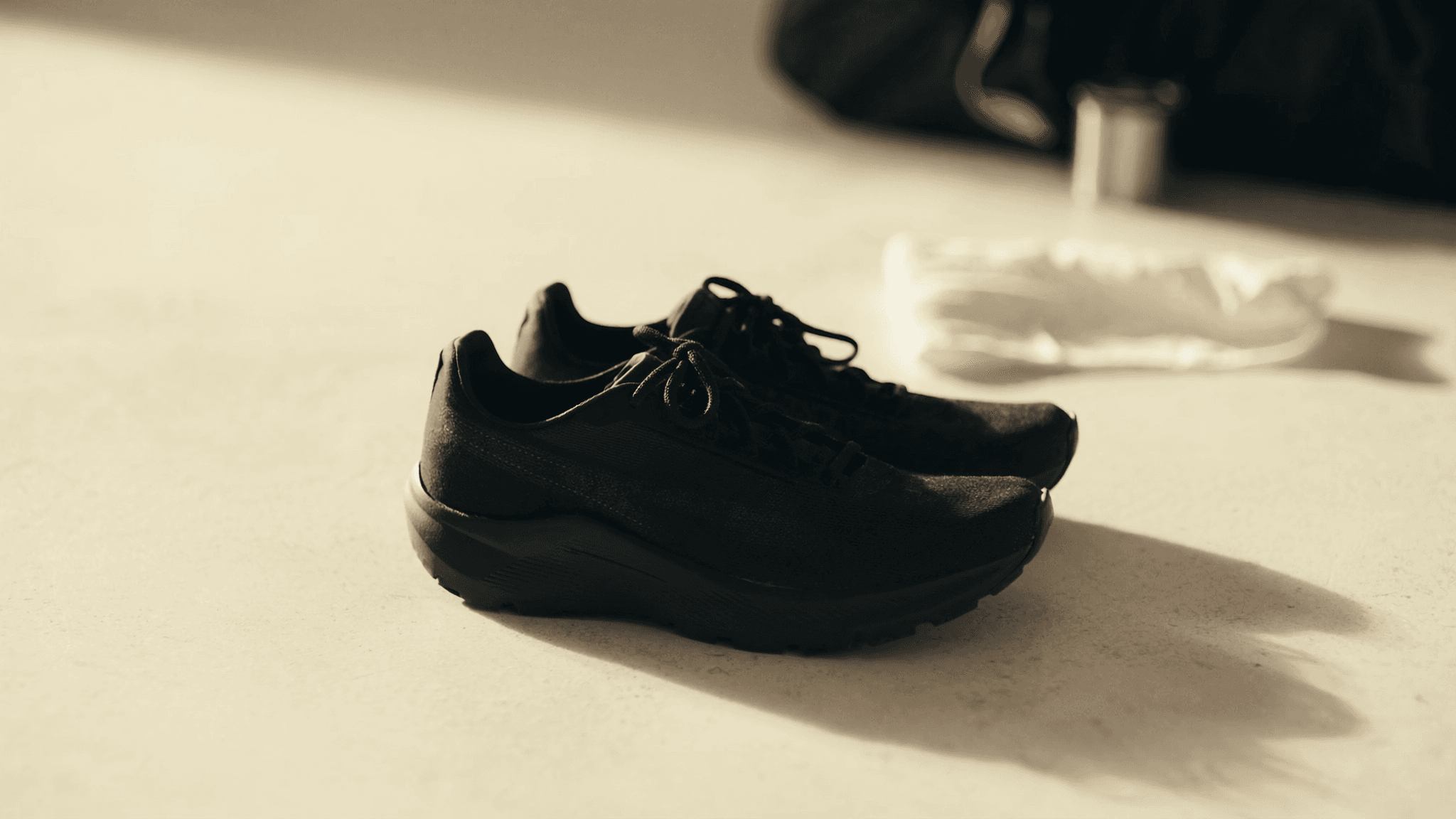 A single pair of matte black minimalist running shoes resting side by side on a warm cream concrete floor, lit by a clean diagonal pool of warm morning daylight from upper-left; a rolled white cotton towel and a charcoal sports kit bag in soft focus background.