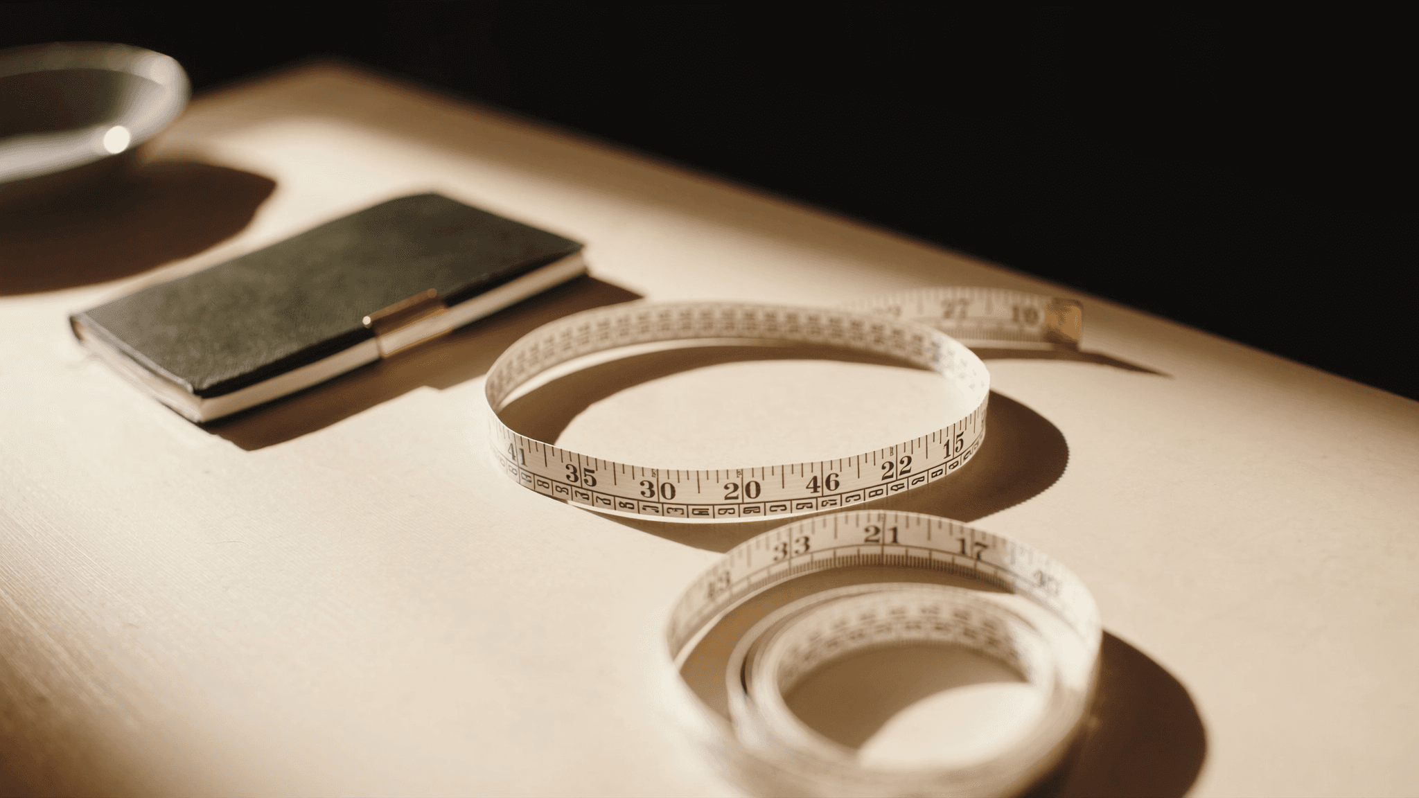 A soft fabric tailor's measuring tape unwound in a graceful curve on a warm cream wooden desk, with measurement markings visible in black numerals; a charcoal leather notebook and a small ceramic dish in soft focus in the background.