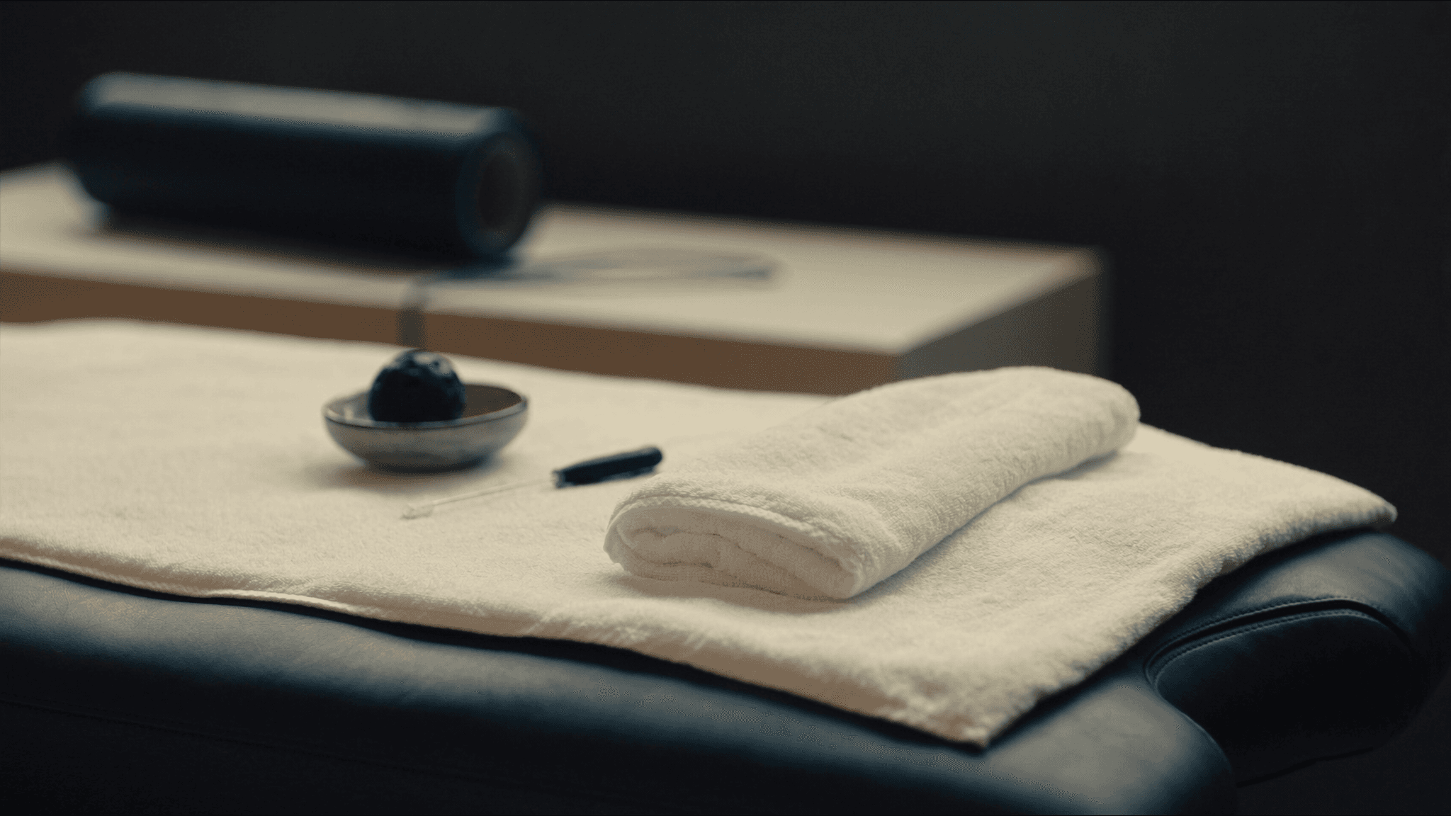 A clinical soft-tissue therapy setup in a premium private training studio: matte black leather massage table, folded white cotton towel as the visual anchor, a small ceramic dish containing a black lacrosse ball and a slim trigger-point cane, with a foam roller resting on a low credenza in soft focus behind