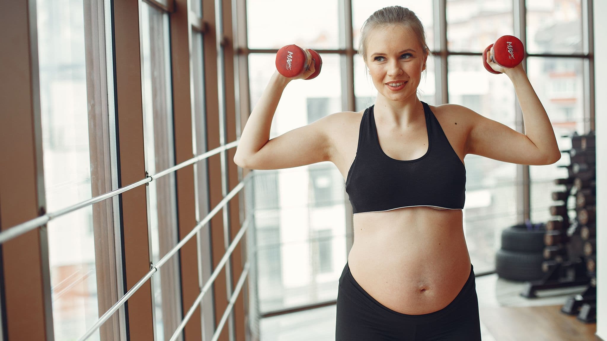 A pregnant woman lifting dumbbells overhead during a gym workout
