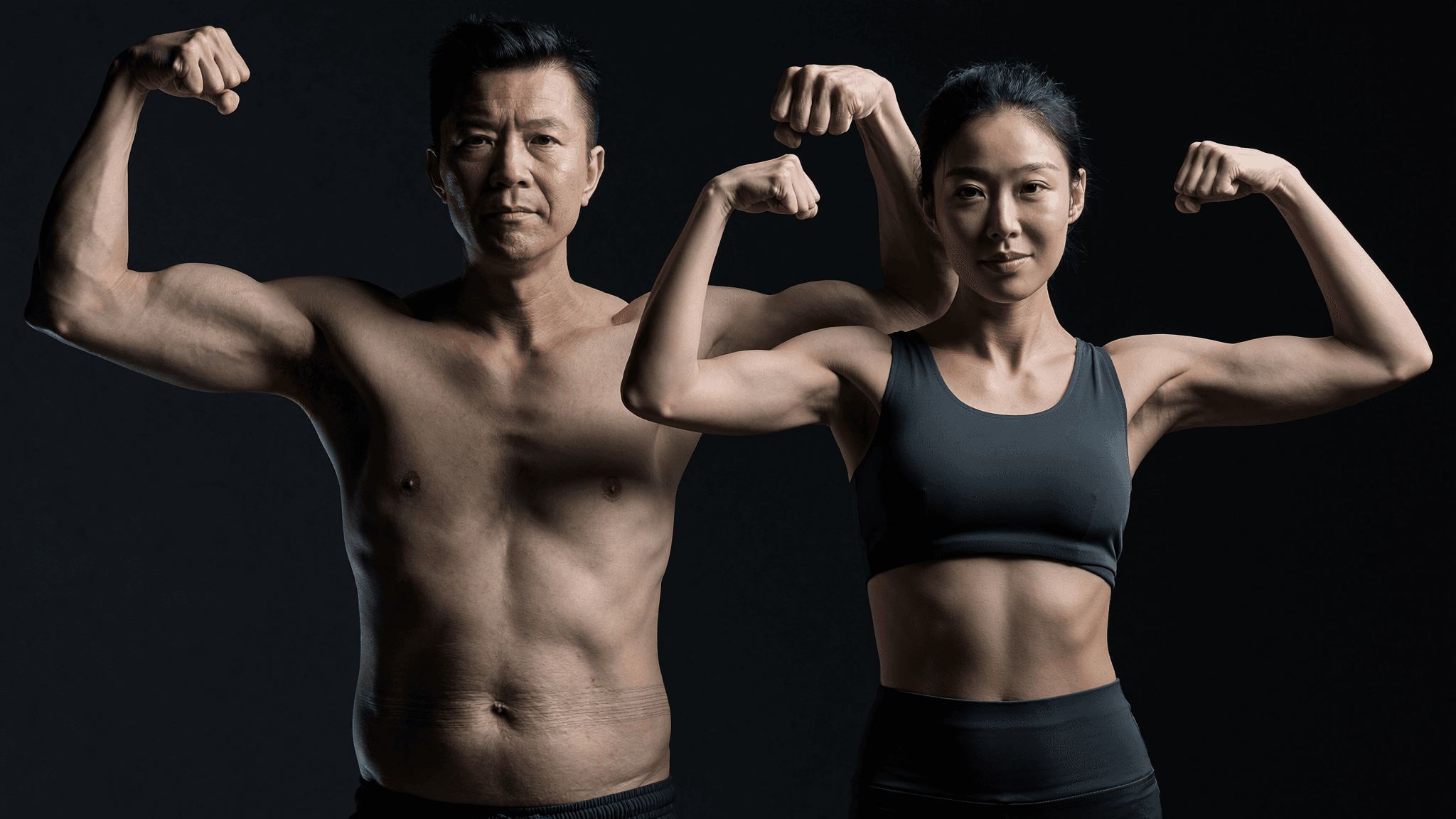 An Asian husband and wife in their late 40s holding the classic front double biceps transformation pose. Both visibly lean, both visibly real: subtle loose-skin texture on his lower abdomen from major weight loss, soft postpartum midsection on hers. The honest version of the aesthetic the transformation-gym market sells.